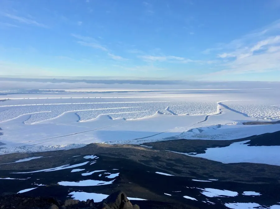 A person in cold weather gear standing on an icy hill overlooking a body of water.
